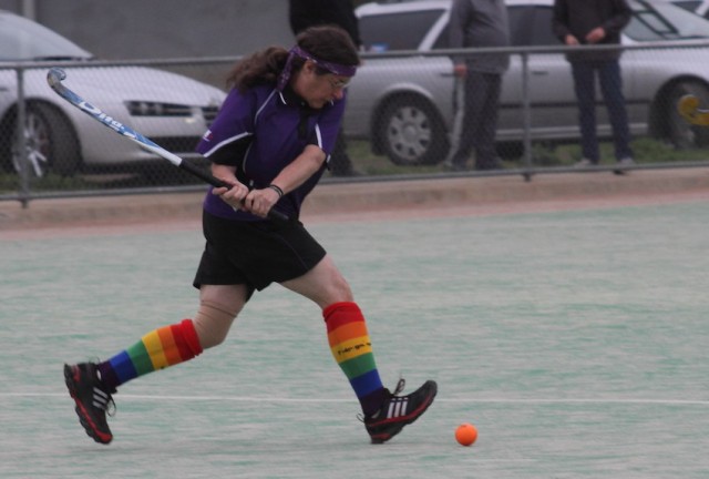 Sandy is on a hockey pitch with her hockey stick in position to hit a ball. She is wearing a purple uniform and rainbow coloured socks.