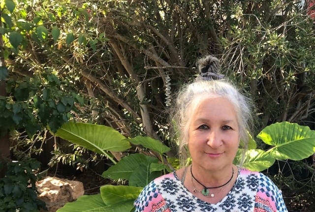 Sandra is standing outside, smiling and wearing a patterned shirt. Sun is shining on her and the trees and plants behind her.