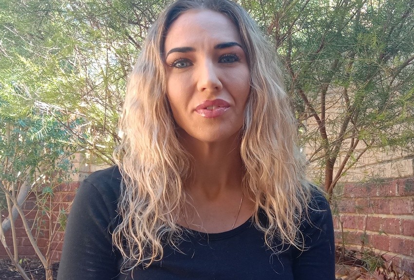 Person from shoulders up with long blond hair smiling at camera. They are outside and there are trees in the background.