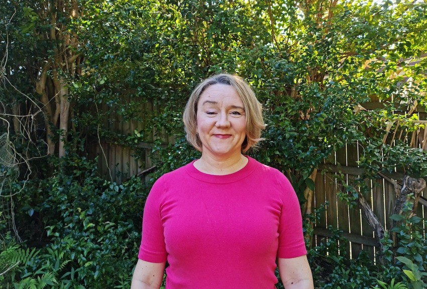 Anita wearing bright pink and standing outside in front of a garden fence covered with vines