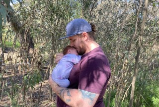 Hamish holds Matilda to his chest and kisses her head there are trees behind them