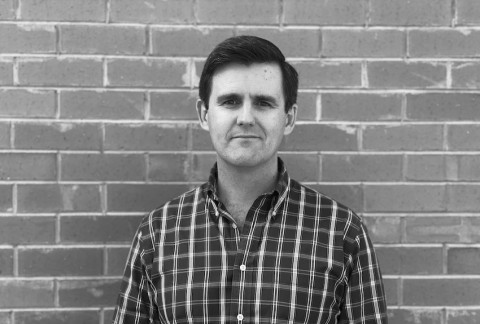 Nick is looking into the camera with a small smile, wearing a checked shirt and there is a brick wall behind him. 