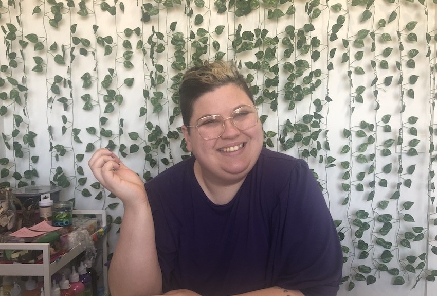 Jess smiles sitting at a workstation with art supplies next to them and vines decorating the wall behind them.