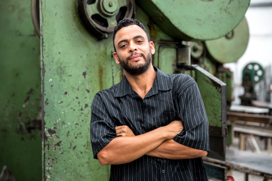 Man with a short beard leaning aginst a wall with his arms folded