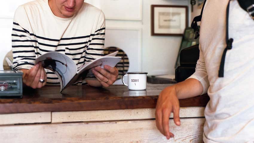 One man is reading with a coffee beside him and another man leans on the table to chat