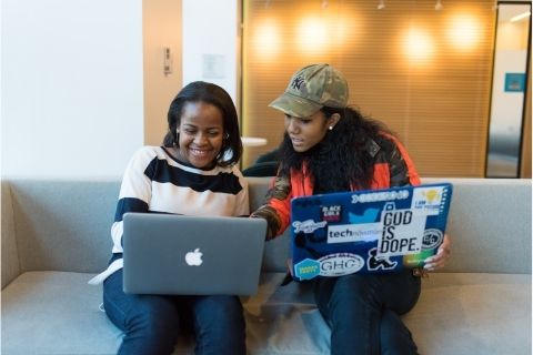 Two people sitting with laptops, one person is smiling, another is pointing at their screen