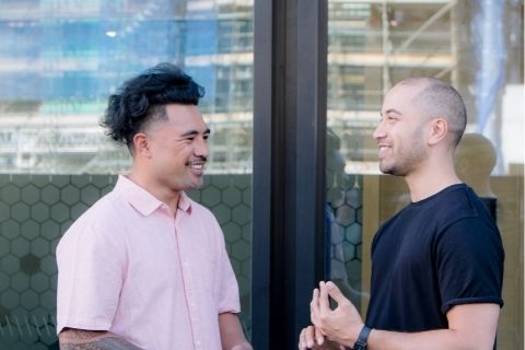 Two people chat outside of a building they are smiling and one is gesturing with their hands