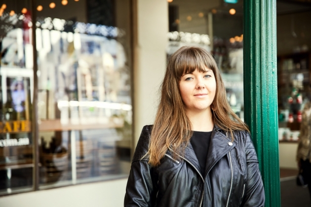 Elly, a young woman with long hair wearing a black leather jacket, standing in front of a shop