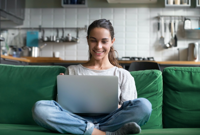 Person sits cross legged on their couch smiling at a computer in their lap